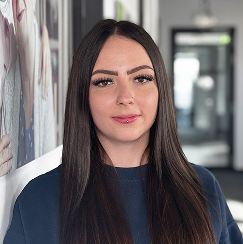 Eine Frau mit langen glatten dunklen Haaren und einem Nasenpiercing steht in einem Haus, blickt in die Kamera und lächelt leicht. Sie trägt ein dunkles Oberteil, und im Hintergrund ist ein verschwommener Flur zu sehen.
