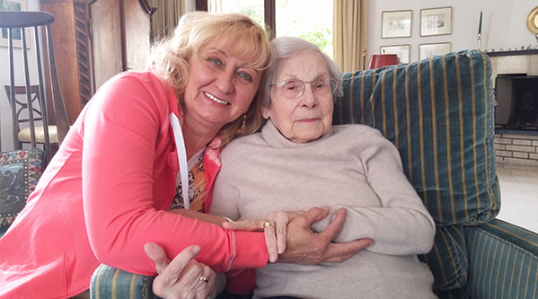 Eine lächelnde Frau mit blondem Haar umarmt eine ältere Frau mit grauem Haar und Brille, die in einem gestreiften Sessel sitzt. Sie befinden sich in einem gemütlichen Wohnzimmer und genießen die warme Unterstützung durch eine 24-Stunden-Betreuungskraft.