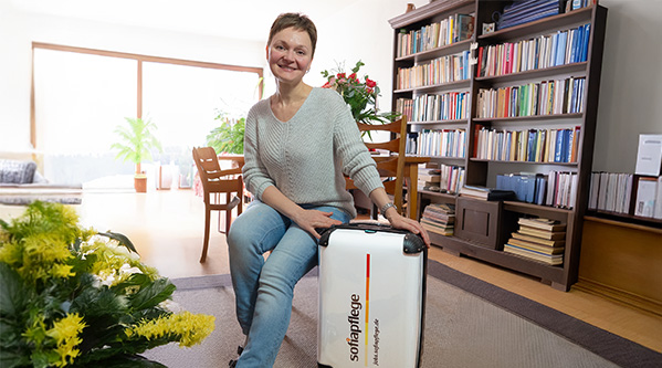 Eine lächelnde Betreuungskraft sitzt auf einer Couch in einem hellen Wohnzimmer und stützt ihre Hand auf einen weißen Koffer mit der Aufschrift sofapflege - perfekt für alle, die 24-Stunden-Pflege suchen. Bücherregale und Blumen schaffen eine gemütliche, einladende Atmosphäre.