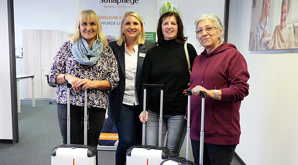 3 Betreuungskräfte und eine Fachberaterin stehen in einem Raum und lächeln in die Kamera, jede hält den Griff eines weißen Koffers. In einem hellen Büro mit Plakaten und Pflanzen im Hintergrund repräsentieren sie die engagierten Dienste 24-Stunden-Pflege und 24-Stunden-Betreuungskraft.