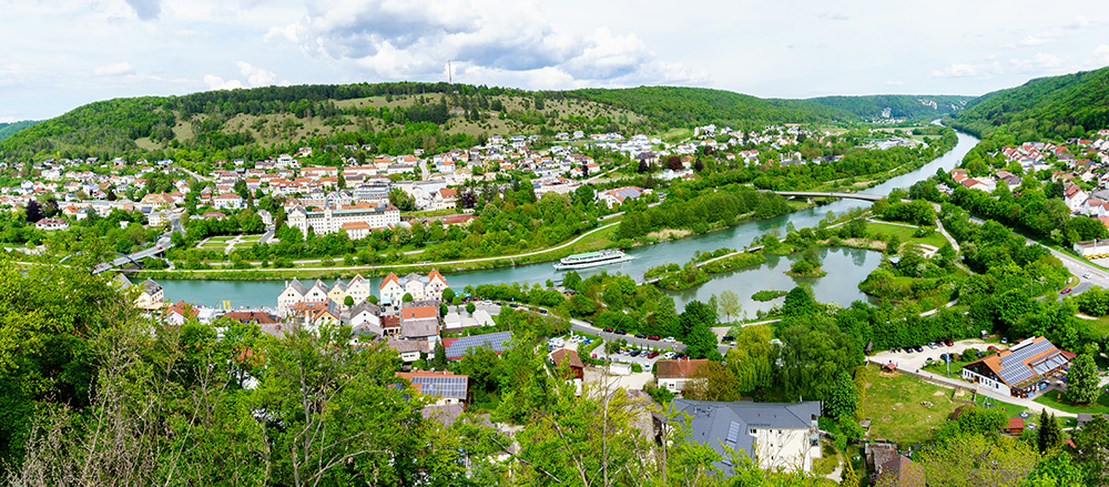 Ein Panoramablick auf eine Stadt mit roten Dächern, eingebettet zwischen grünen Hügeln in der Hajo Penther - Sofiapflege Region Nordsee. Ein gewundener Fluss, wahrscheinlich die Elbe oder Weser, fließt durch die Landschaft mit Brücken und einem Boot unter einem teilweise bewölkten Himmel.