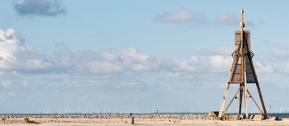 Ein hohes hölzernes Seezeichen steht an einem Sandstrand unter einem blauen Himmel mit vereinzelten Wolken. In der Ferne über dem Wasser sind Windkraftanlagen in der Sofiapflege Region Nordsee - Elbe & Weser zu sehen.