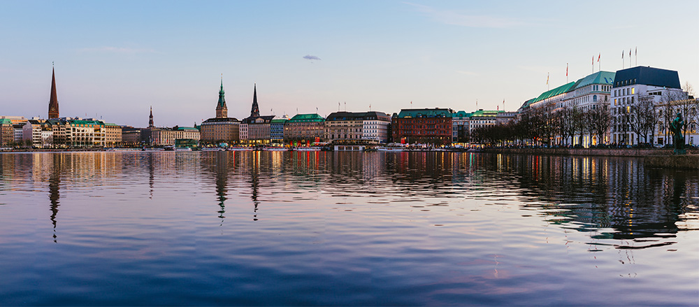 Panoramablick auf Gebäude und Kirchtürme am Ufer der Alster in Hamburg in der Abenddämmerung, mit Spiegelungen im Wasser und einem klaren Himmel darüber - eine Aufnahme der ruhigen Schönheit bei Oliver Mielchen - Sofiapflege Schleswig-Holstein & Oberalster.