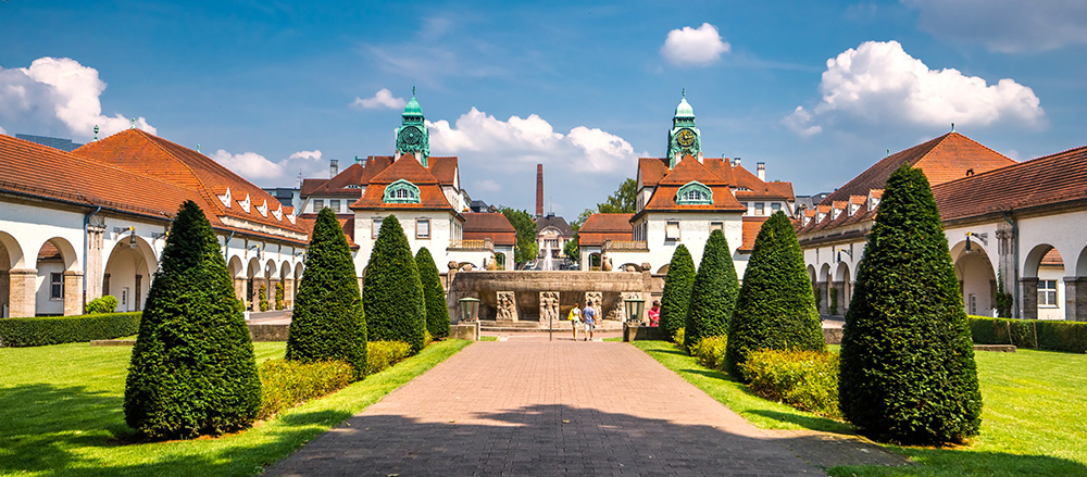 Ein gepflegter Garten mit ordentlich geschnittenen kegelförmigen Büschen führt zu einem kunstvollen historischen Gebäude von Heiko Beck mit zwei Uhrentürmen, roten Ziegeldächern und weißen Wänden unter einem sonnigen blauen Himmel mit vereinzelten Wolken.