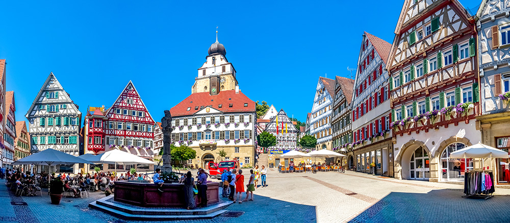 Ein lebendiger Stadtplatz in Herrenberg mit farbenfrohen, traditionellen Fachwerkhäusern, Straßencafés mit Sonnenschirmen, Menschen, die spazieren gehen und sitzen, ein zentraler Brunnen und eine historische Kirche unter einem klaren blauen Himmel.