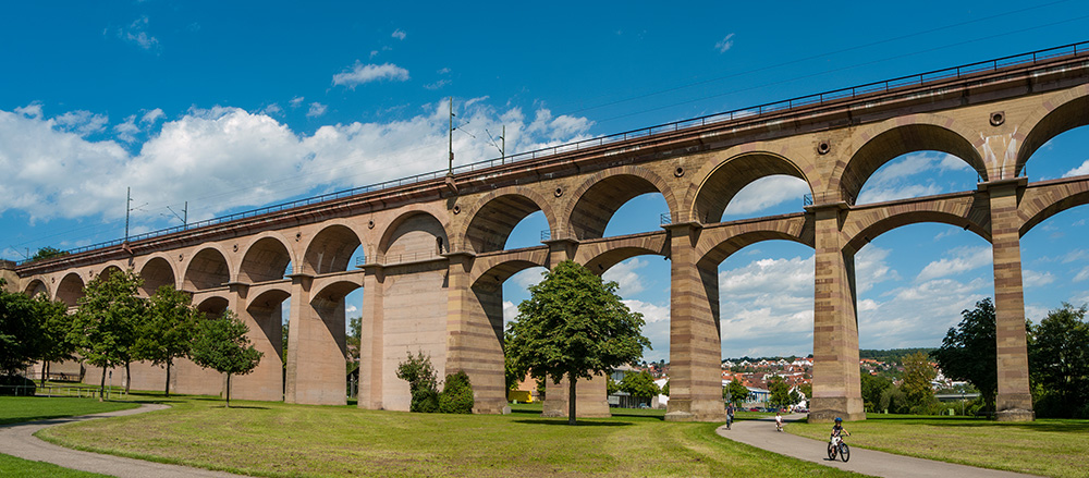 Ein großes steinernes Viadukt in Bietigheim-Bissingen mit mehreren Bögen überspannt einen grasbewachsenen Park mit Bäumen und einem Radfahrer auf einem Weg darunter. Der Himmel ist blau, mit vereinzelten Wolken.
