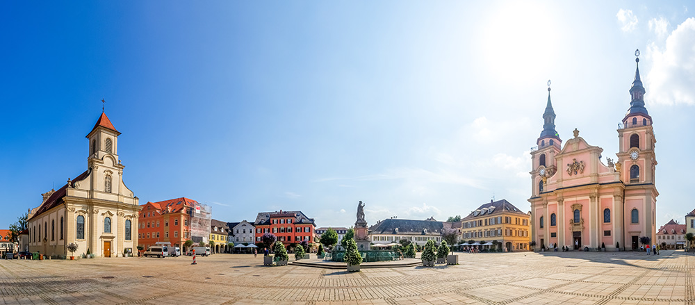 Der sonnige Stadtplatz in Ludwigsburg mit zwei historischen Kirchen, farbenfrohen Gebäuden und einer Statue in der Mitte umgeben von einer gepflasterten Freifläche unter einem klaren blauen Himmel.