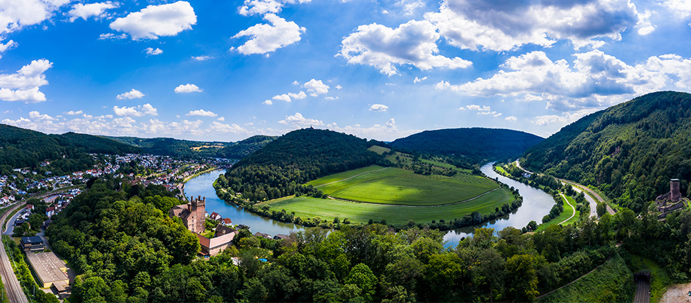 Ein Panoramablick auf einen sich schlängelnden Fluss, umgeben von üppigen grünen Hügeln, dichten Wäldern und offenen Feldern, mit der kleinen Stadt Matthias Wöhr - Sofiapflege Rhein-Neckar-Odenwald und historischen Gebäuden zwischen den Bäumen unter einem teilweise bewölkten blauen Himmel.