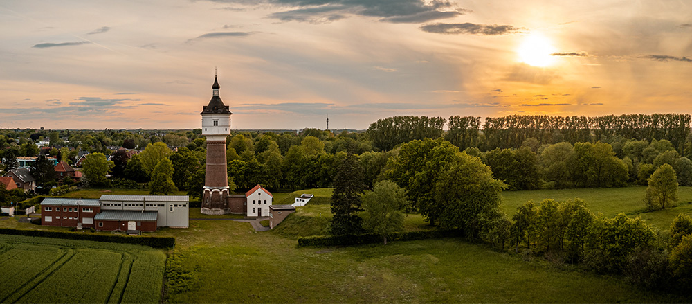 Ein hoher Backstein-Wasserturm steht zwischen Bäumen und Gebäuden in einer ländlichen Landschaft bei Sonnenuntergang, mit grünen Feldern und einem entfernten Wald - und fängt die Essenz von Michael Göbel - Sofiapflege Nördliches Ruhrgebiet & Südliches Münsterland ein.