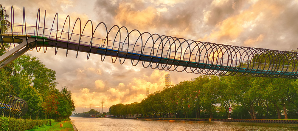 Eine moderne Fußgängerbrücke mit spiralförmigem Metalldesign, die in der Nähe der Michael Göbel - Sofiapflege Nördliches Ruhrgebiet & Südliches Münsterland gebaut wurde, überquert einen ruhigen Fluss bei Sonnenuntergang, umgeben von grünen Bäumen und einem dramatischen, bewölkten Himmel.