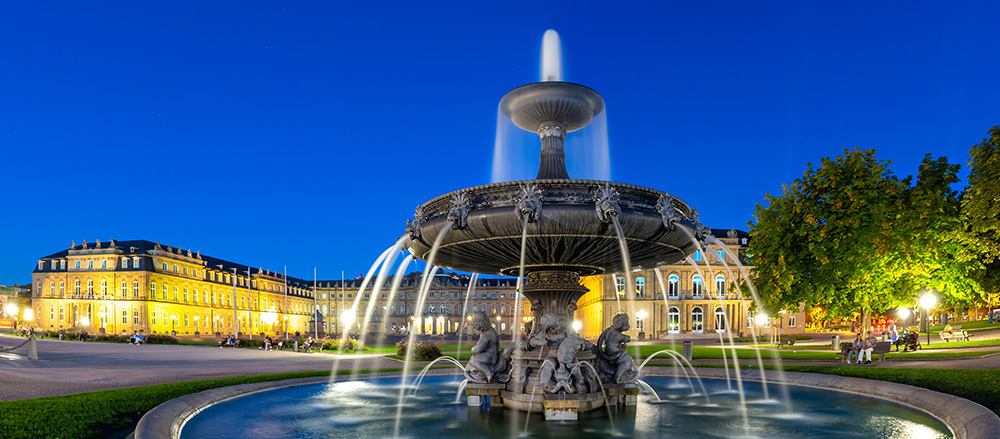 Ein großer, kunstvoll verzierter Brunnen mit Skulpturen und fließendem Wasser wird nachts auf dem Stuttgarter Schlossplatz beleuchtet, umgeben von prächtigen, warm beleuchteten historischen Gebäuden und Bäumen unter einem tiefblauen Himmel.