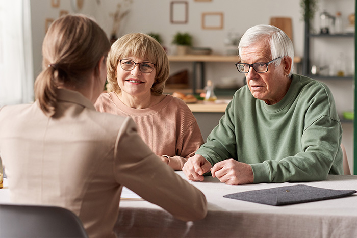 Ein älteres Ehepaar sitzt einer Frau im Blazer an einem Tisch gegenüber und diskutiert über Pflegeberatung nach § 37 Abs. 3 SGB XI in einem hellen, gemütlichen Raum mit Regalen und Dekoration im Hintergrund.