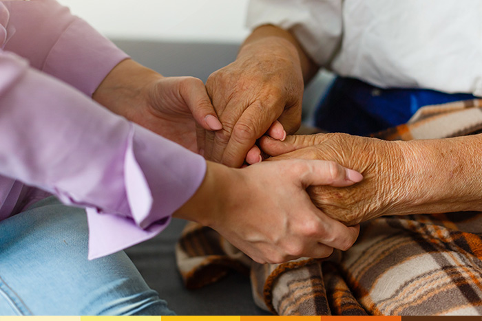 Eine jüngere Person hält sanft die Hände eines älteren Erwachsenen, beide sitzen eng beieinander. Die ältere Person ist mit einer karierten Decke zugedeckt, wodurch eine warme und fürsorgliche Atmosphäre entsteht - ein idealer Moment für die persönliche Pflegeberatung nach § 37 Abs. 3 SGB XI.