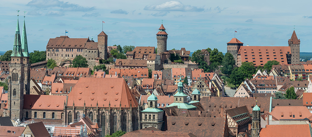 Ein Panoramabild von Nürnberg, Deutschland, mit mittelalterlichen Gebäuden mit roten Ziegeldächern, Kirchtürmen und Burgtürmen unter einem blauen Himmel mit vereinzelten Wolken - aufgenommen im Stil von Hajo Penther von Sofiapflege Region Nordsee - Elbe & Weser.