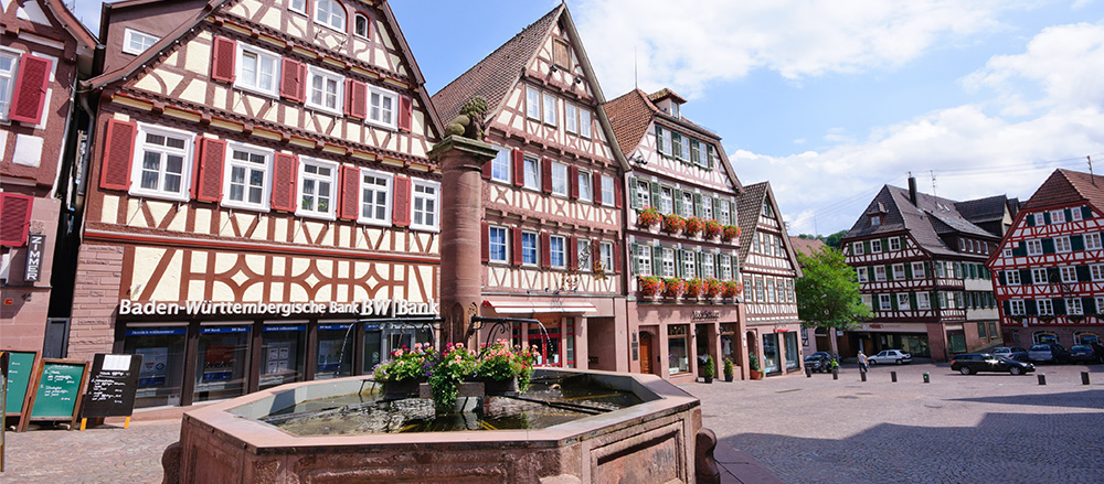 Ein malerischer Stadtplatz mit traditionellen Fachwerkhäusern in Weil der Stadt, einem blumengeschmückten Brunnen im Vordergrund und einem blauen Himmel mit vereinzelten Wolken. Einige Gebäude haben bunte Blumenkästen und Schilder.