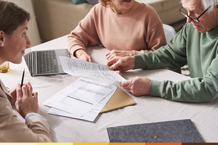 Drei Personen sitzen an einem Tisch und sehen gemeinsam Unterlagen durch, es geht um Pflegeberatung nach § 37 Abs. 3 SGB XI. Einer hält einen Stift in der Hand, ein anderer zeigt auf die Papiere; ein Laptop und ein Briefumschlag deuten auf finanzielle oder pflegebezogene Unterlagen hin.