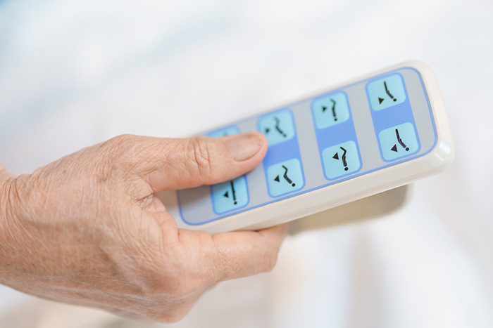Eine Nahaufnahme der Hand einer älteren Person, die eine Fernbedienung mit blauen Tasten hält, die zum Einstellen der Position eines verstellbaren Bettes verwendet wird - was die Bedeutung der Technischen Pflegehilfsmittel-Beratung unterstreicht.