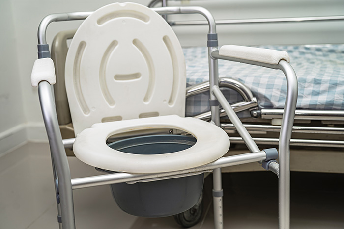 Ein Nachttischstuhl mit weißer Sitzfläche und Metallgestell steht neben einem Krankenhausbett mit einer blau-weiß karierten Decke, ideal für Menschen, die Technische Pflegehilfsmittel Beratung suchen.