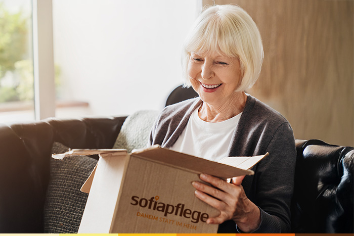 Eine ältere Frau mit grauen Haaren öffnet lächelnd einen Karton mit der Aufschrift sofiapflege, während sie auf einer Couch in einem hellen, gemütlichen Wohnzimmer sitzt.
