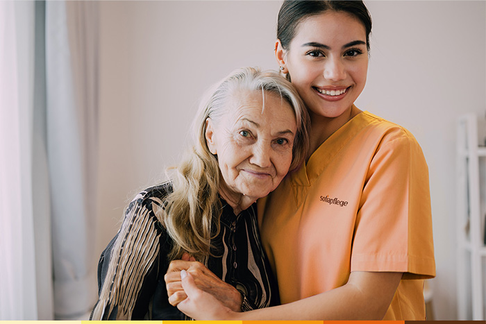 Eine lächelnde ältere Frau wird von einer jungen Pflegerin in orangefarbenem Kittel umarmt. Sie stehen eng beieinander in einem gut beleuchteten Raum und blicken beide mit warmem Gesichtsausdruck in die Kamera.