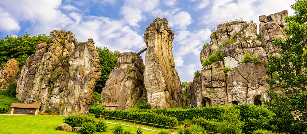 Hohe, schroffe Felsformationen erheben sich über grüne Sträucher und Grasflächen unter einem teilweise bewölkten blauen Himmel im teuteborger Wald. Am Fuße der Felsen stehen kleine Holzhäuser, die an die beschauliche 24-Stunden-Pflege in Osnabrück erinnern.