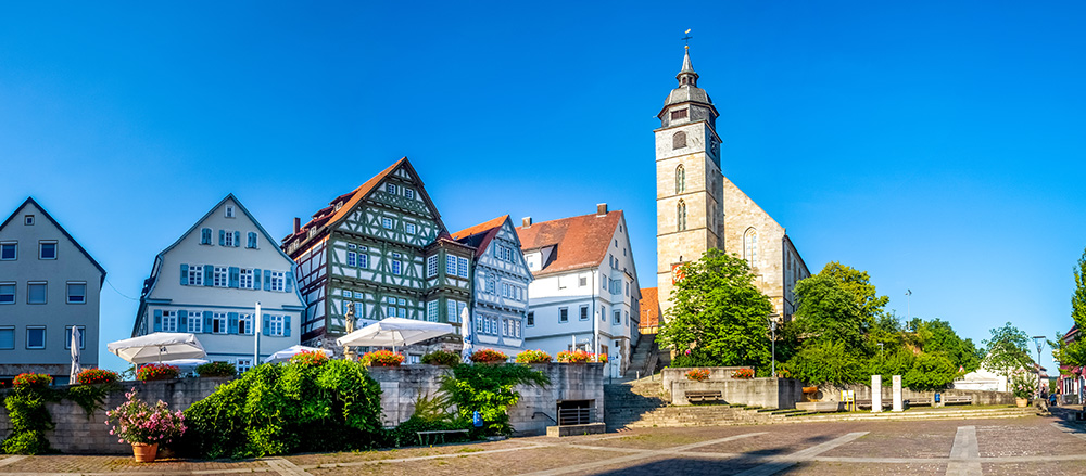 Stadtplatz Böblingen mit Fachwerkhäusern, Sitzgelegenheiten im Freien mit Sonnenschirmen, Blumenkübeln und einem hohen steinernen Kirchturm unter einem klaren blauen Himmel. Bäume säumen den Platz und sorgen für eine grüne Kulisse.