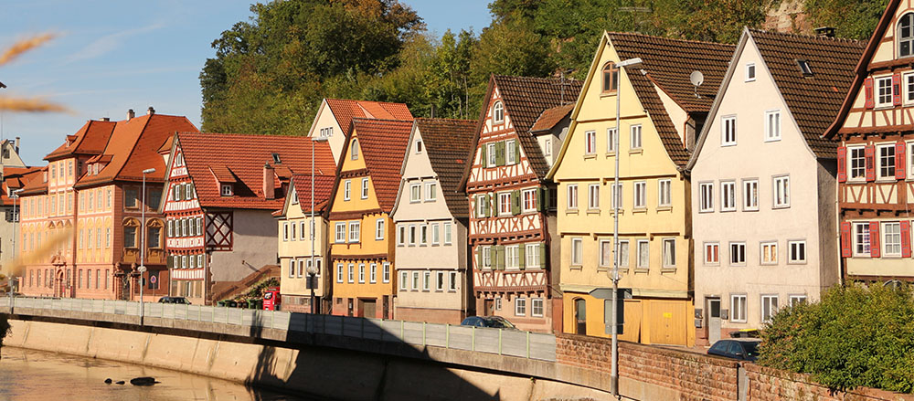 Eine Reihe farbenfroher, traditioneller Fachwerkhäuser mit steilen Dächern säumt eine Straße am Flussufer unter einem klaren blauen Himmel, mit Bäumen im Hintergrund.