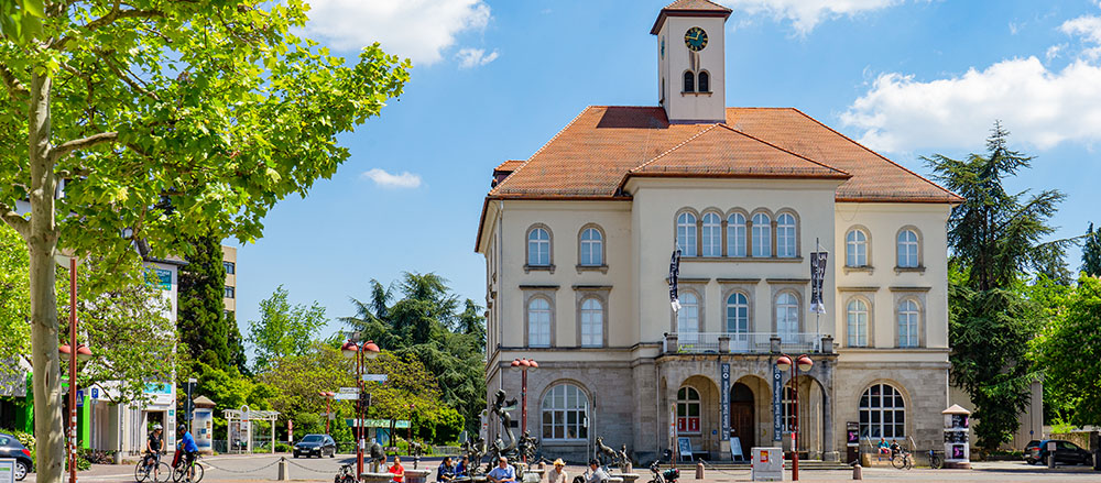 Ein großes, historisches Gebäude in Sindelfingen mit einem Uhrenturm steht unter blauem Himmel. Davor spazieren und radeln die Menschen, rund um den belebten Stadtplatz sind Bäume und Straßenschilder zu sehen.