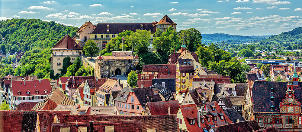 Eine malerische Ansicht von Tübingen, mit bunten Häusern mit Ziegeldächern, gewundenen Straßen, üppigen grünen Bäumen und einem großen schlossähnlichen Gebäude auf einem Hügel unter einem blauen Himmel mit vereinzelten Wolken.