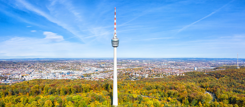 Ein hoher Fernmeldeturm erhebt sich über einem Wald mit Herbstlaub und überblickt eine weitläufige Stadt unter einem blauen Himmel mit Wolkenfetzen und fernen Hügeln.