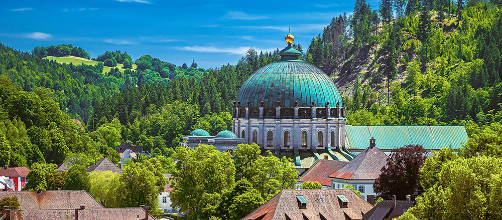 Ein großes Kuppelgebäude in St. Blasien mit grünem Dach steht inmitten von Bäumen und Hügeln, umgeben von kleineren Gebäuden unter einem klaren blauen Himmel in einer üppigen, bewaldeten Landschaft.