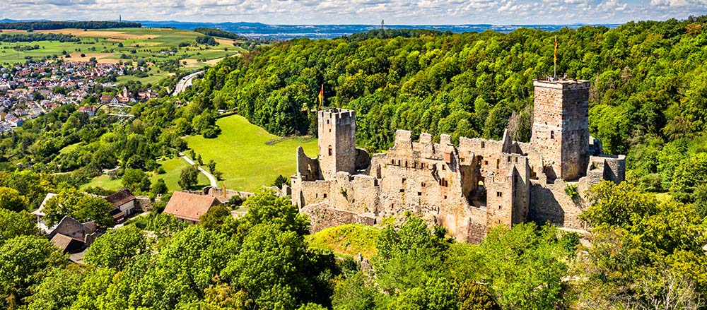 Luftaufnahme einer mittelalterlichen Steinburg in Lörrach und Umgebung, dem Versorgungsgebiet von Simon Gsell, umgeben von einem dichten grünen Wald, mit einem nahe gelegenen Dorf und offenen Feldern unter einem blauen Himmel mit vereinzelten Wolken.