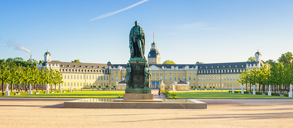 Ein großer gelber Barockpalast in Karlsruhe mit einem zentralen Turm, umgeben von gepflegten Gärten und Bäumen. Im Vordergrund steht eine große Bronzestatue auf einem Sockel, unter einem klaren blauen Himmel.