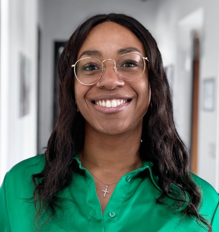 Sara Koppenhöfer, eine lächelnde Frau mit langen dunklen Haaren, die eine Brille und eine grüne Bluse trägt, steht in einem Innenraum. Sie trägt einen Nasenring und eine Kreuzkette. Im Hintergrund ist ein heller, verschwommener Hausflur zu sehen.