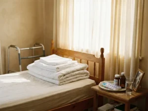 Seniorenschlafzimmer mit frisch gefalteter weißer Bettwäsche, Gehhilfe an der Wand und Medikamenten auf dem Nachttisch im Morgenlicht.