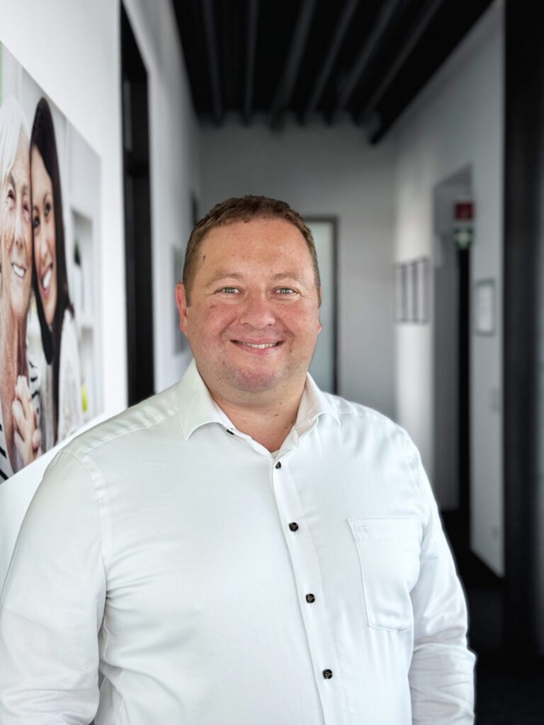 Ein Mann in einem weißen Hemd lächelt in die Kamera; er steht in einem Flur, an dessen Wand neben ihm ein Foto von zwei lächelnden Frauen hängt. Der Hintergrund ist leicht unscharf.